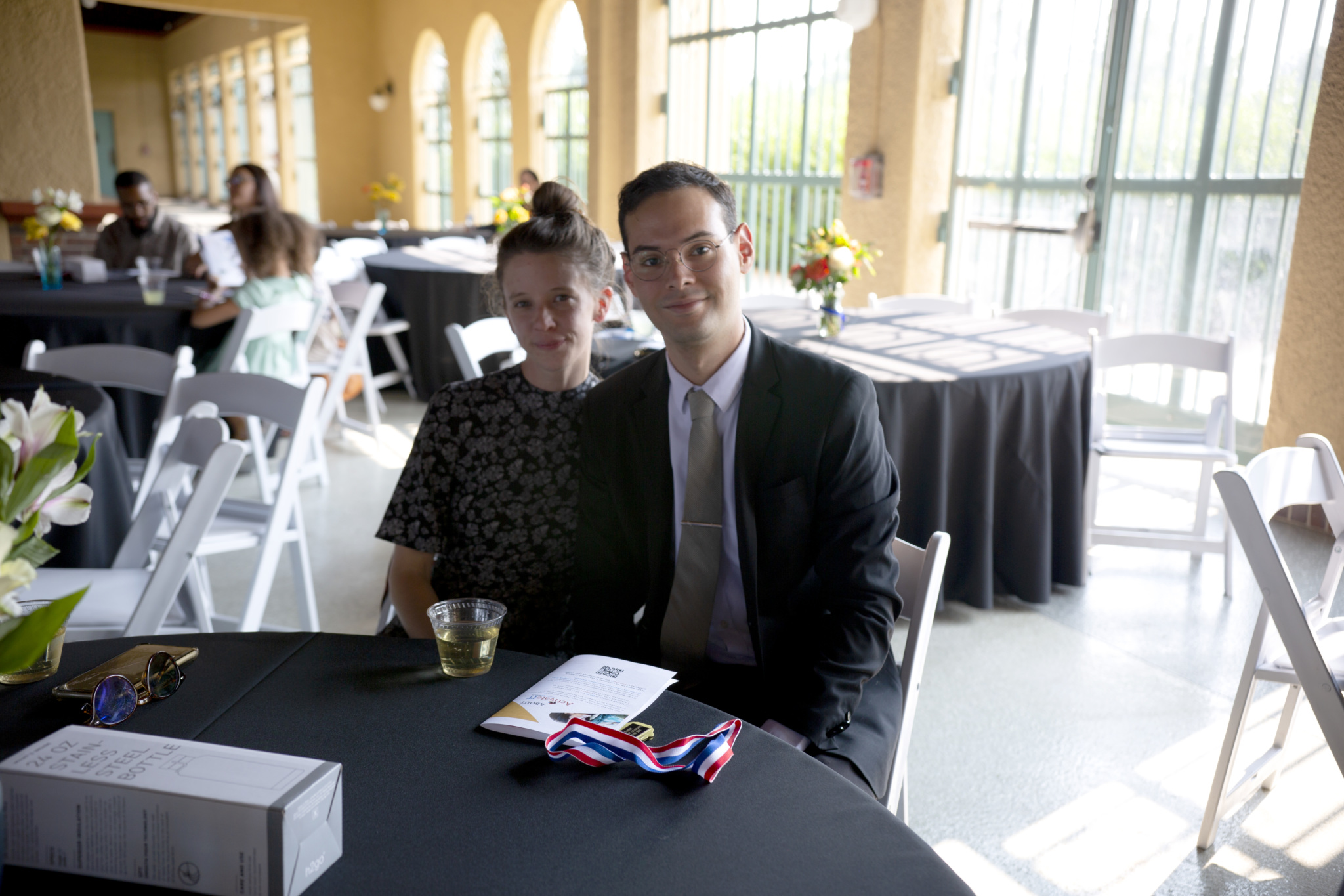 Man and woman seated at a reception table with a program and medals, smiling toward the camera.