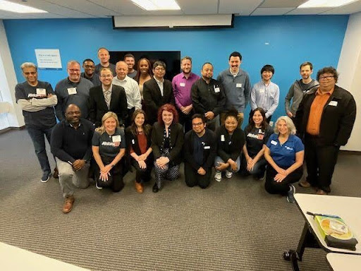 Group of about 24 adults smiling and posing for a group photo in a training room with a blue wall.