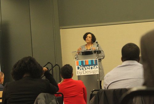 Woman speaking to an audience at a podium labeled "Diverse by Design."