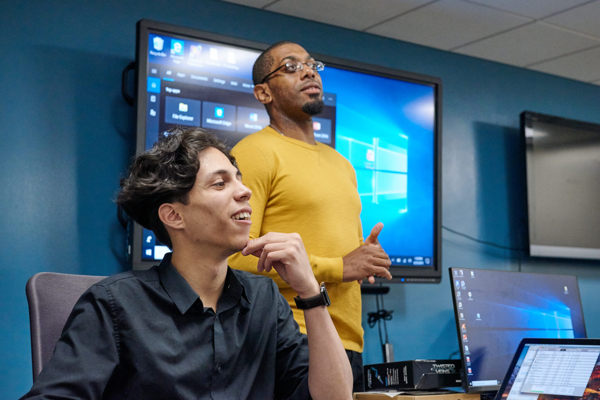 Two learners in a classroom listening and smiling as one stands in front of a large screen displaying a Windows desktop.