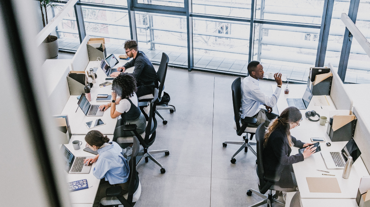 Open-plan office with six young professionals at individual desks working on laptops and devices.