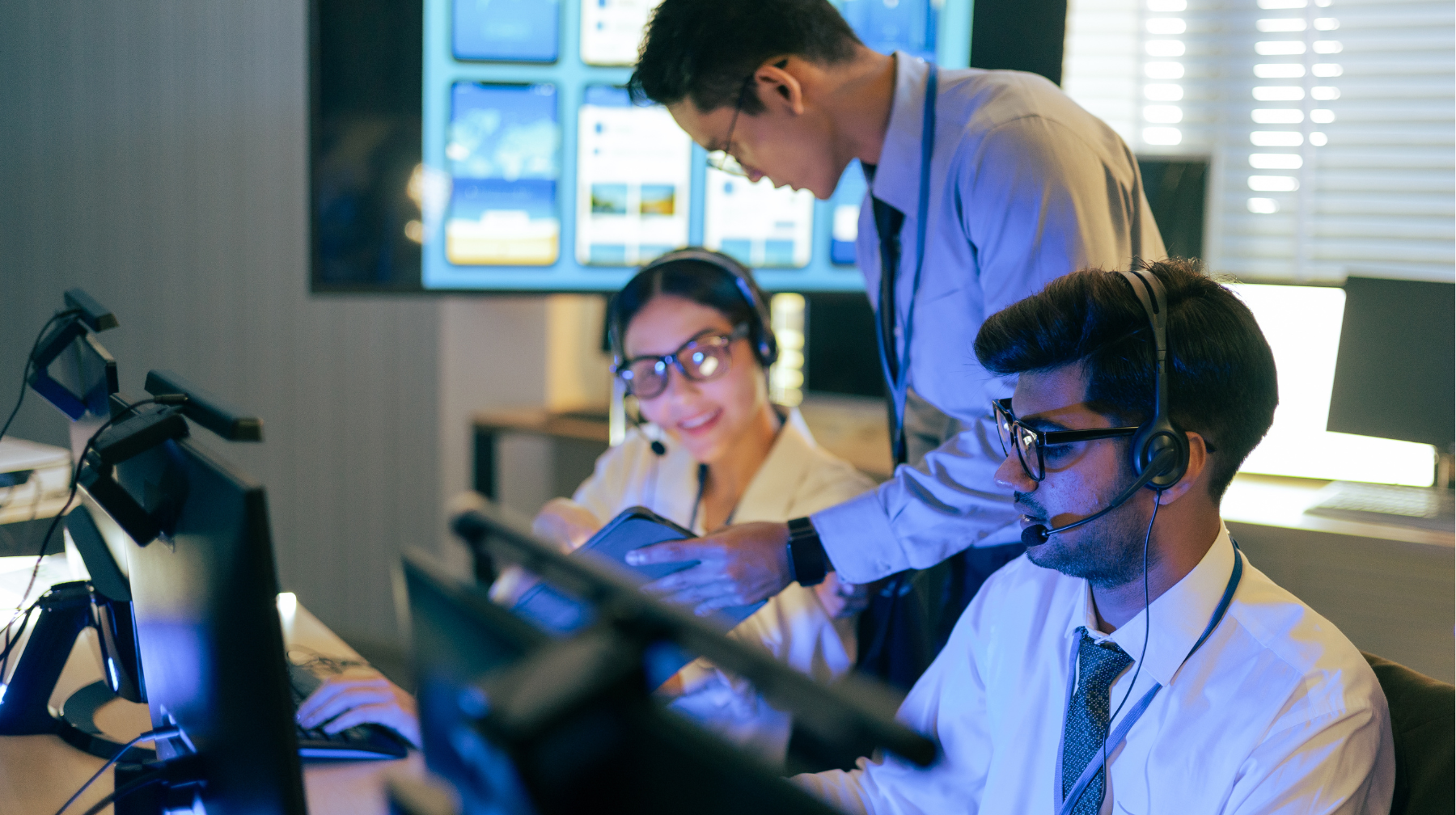 Team of colleagues wearing headsets collaborating at computer monitors with data dashboards displayed on wall screens.
