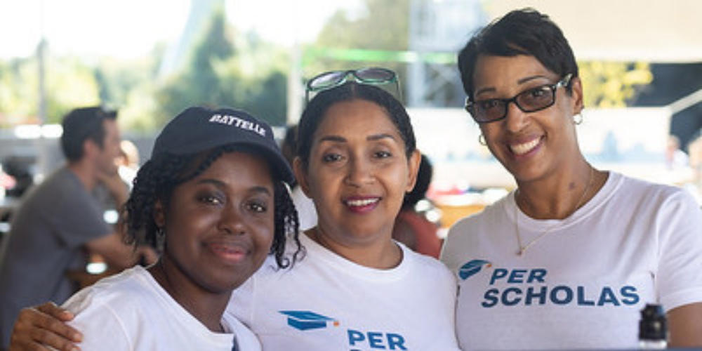 Three smiling adults wearing Per Scholas t-shirts, posing together at an outdoor event.