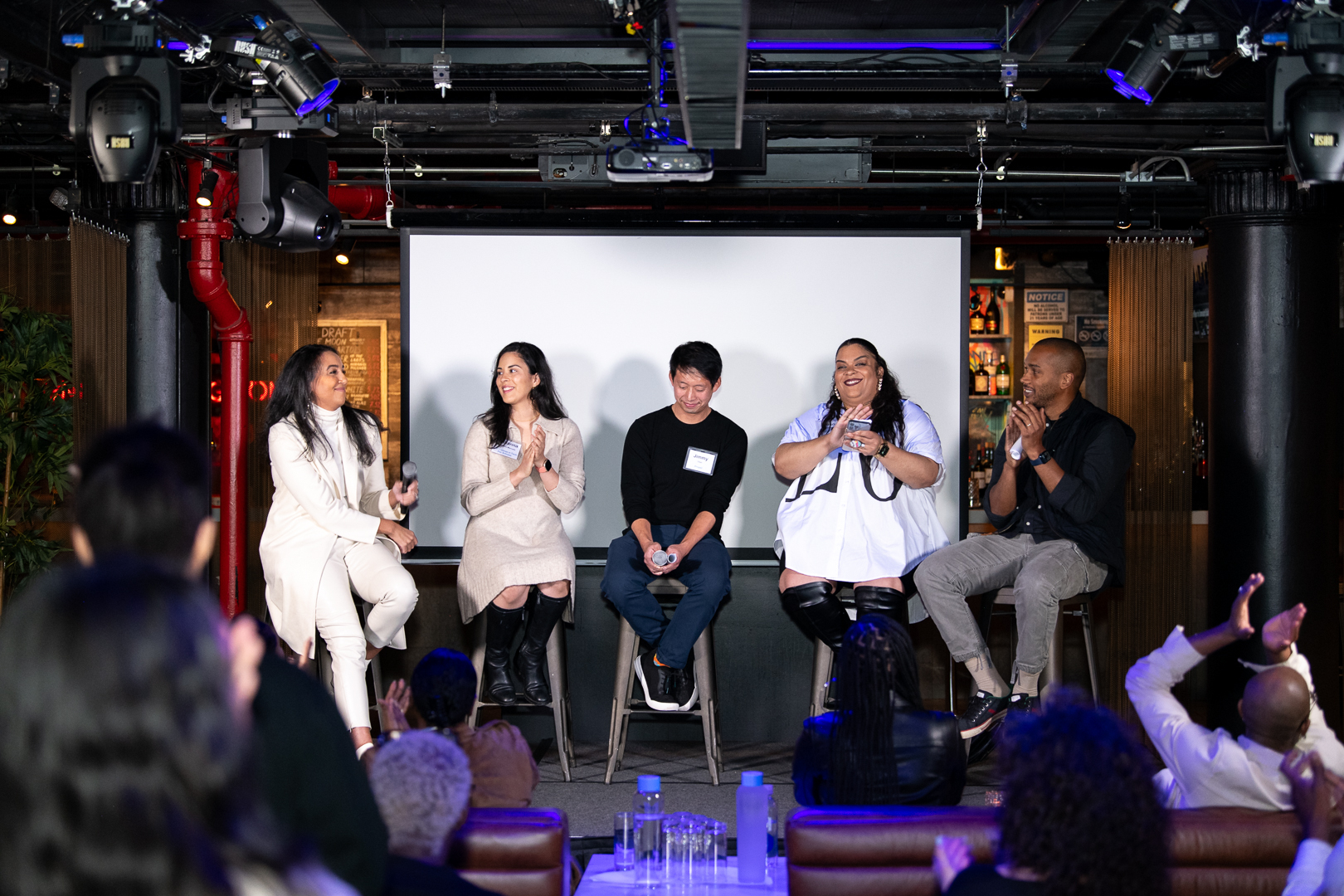Five panelists sit on stools on stage speaking and laughing while an audience watches and applauds.