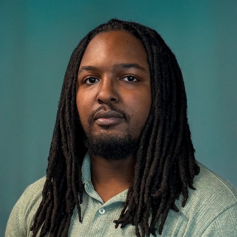 Headshot of a man with long dreadlocks against a plain teal background.