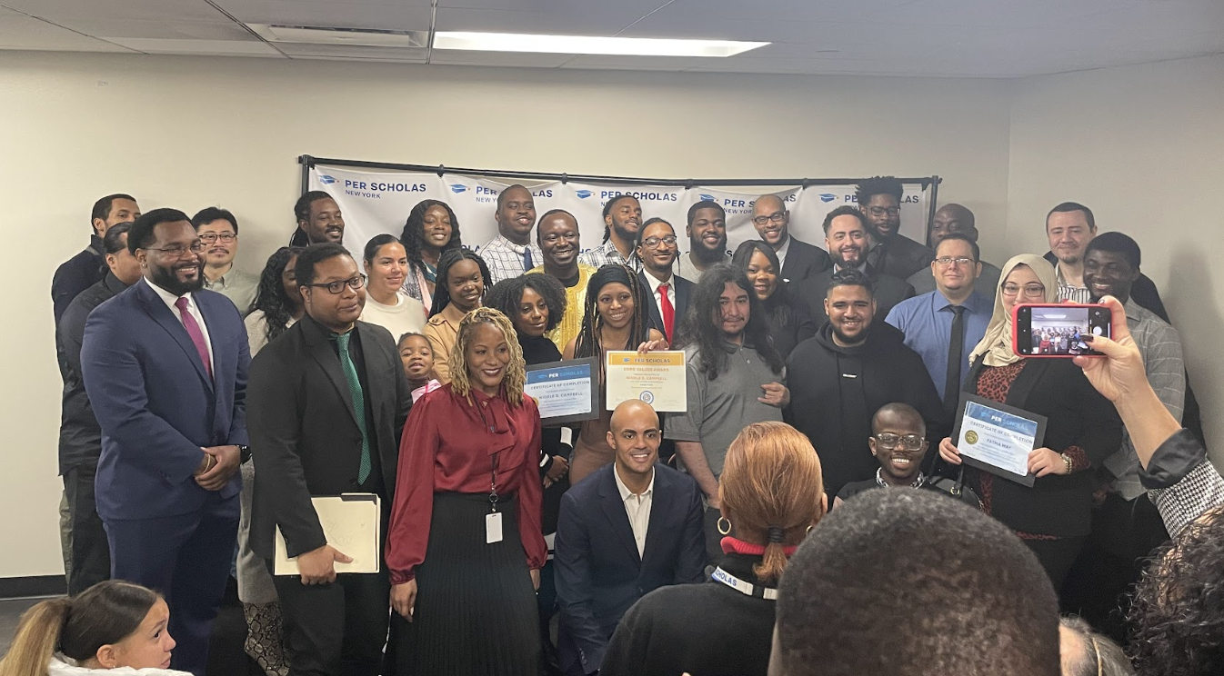 A diverse group of adult program graduates and staff posing for a celebratory group photo with people holding certificates and a Per Scholas banner behind them.