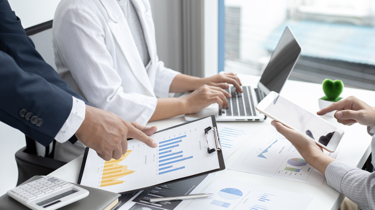 Three people collaborate at a desk, reviewing charts and graphs on printouts, a clipboard, a tablet, and a laptop.