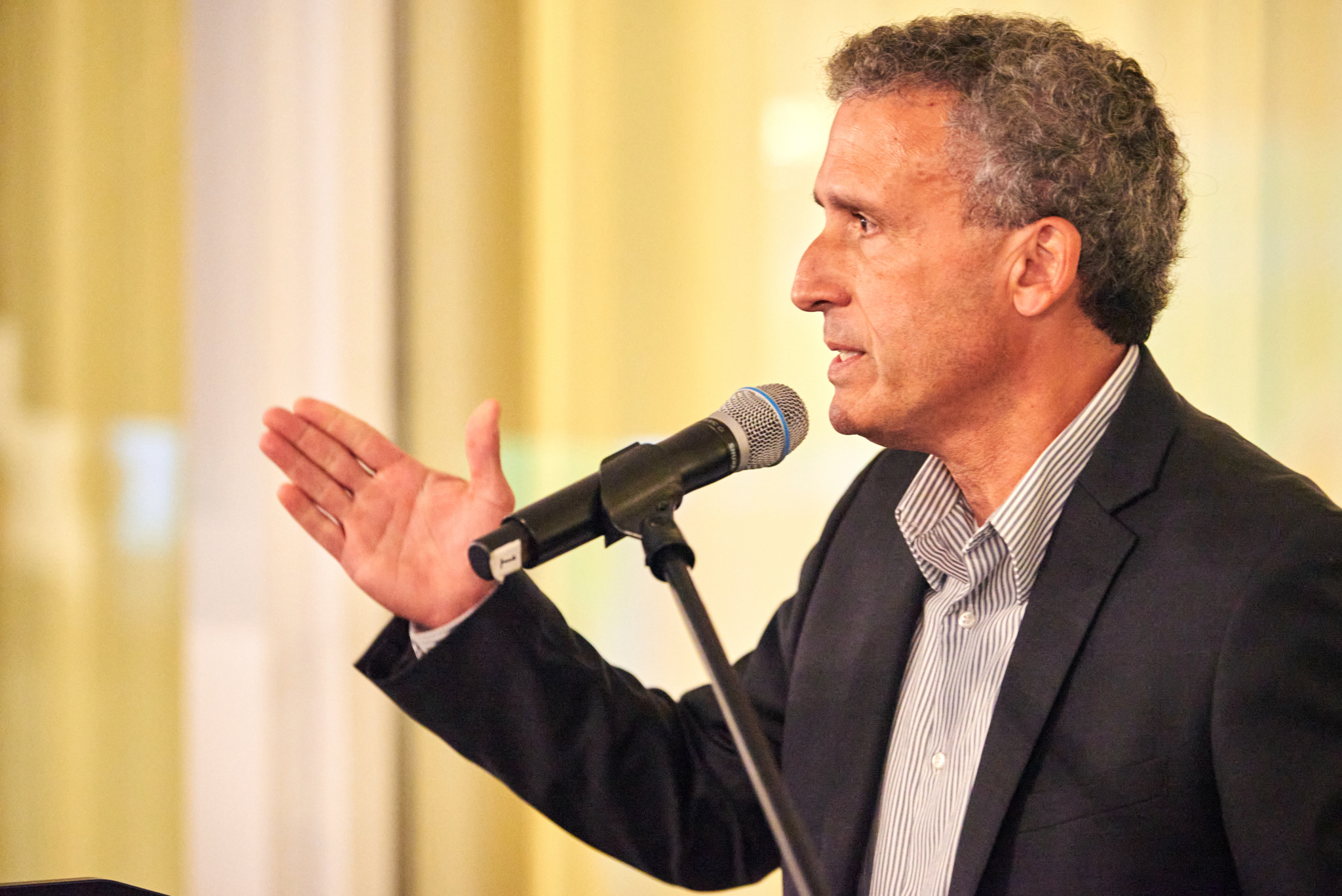 Man speaking into a microphone at a podium, gesturing with his hand as he addresses an audience.
