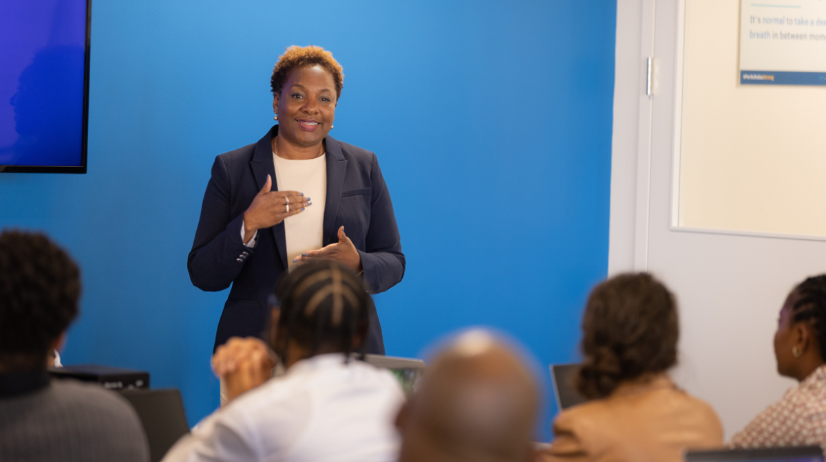 Instructor smiling and teaching a small classroom, speaking to seated students.