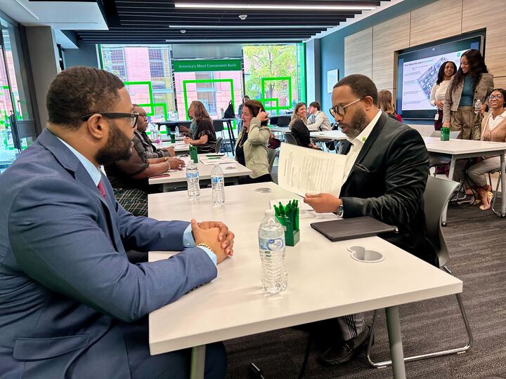 Two men in business suits reviewing a document together at a networking event, with other attendees in conversation in the background.