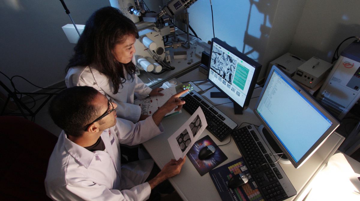 Two lab researchers in white coats review microscope images on printouts and computer monitors.