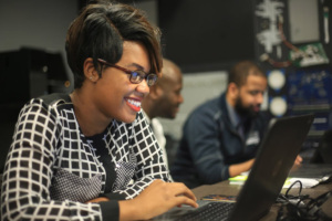Woman student smiling while working on a laptop in a classroom with other learners.