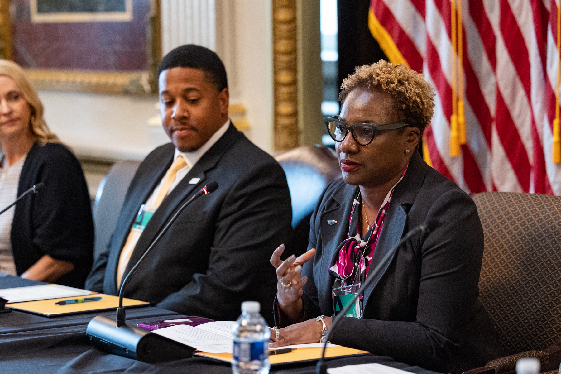 Three people seated at a conference table, with a woman speaking into a microphone while a man and woman listen.