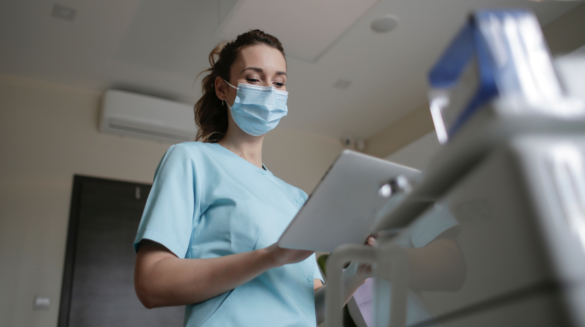 Female healthcare worker wearing a mask and scrubs checks a tablet while standing next to medical equipment.