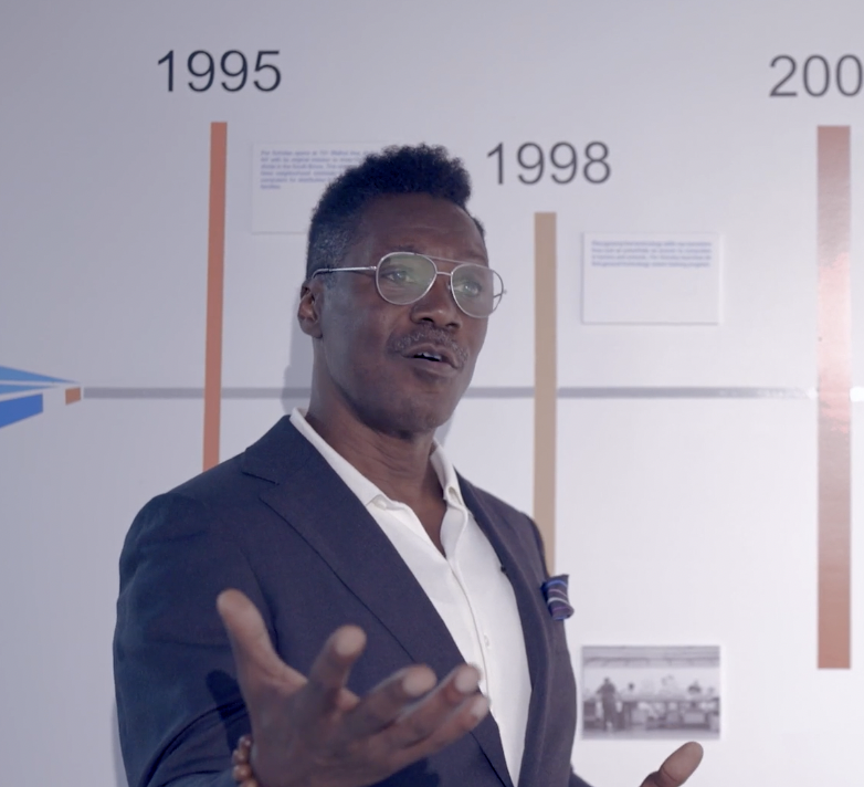 Man speaking and gesturing in front of a timeline wall labeled with years including 1995 and 1998.