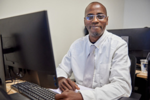 Smiling adult man sitting at a desk working on a desktop computer in an office or classroom setting.