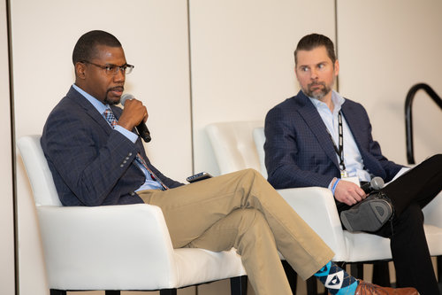 Two men seated on stage in a panel discussion; the left man speaks into a microphone while the right man listens.