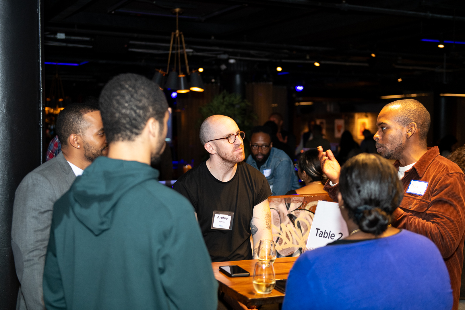 A small group of adults standing and sitting around a table having a conversation at a networking event.