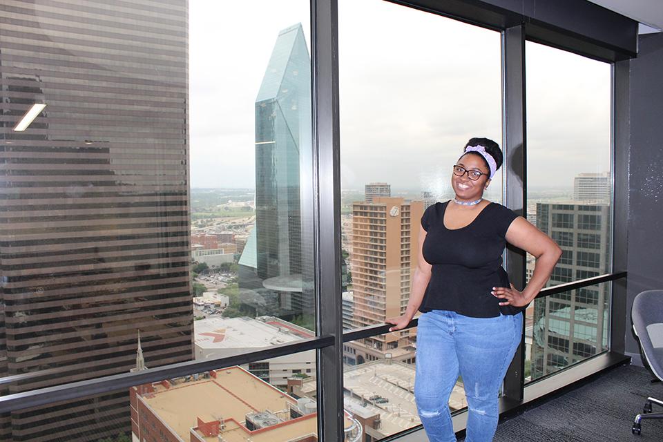 Young woman smiling and posing by a high-rise window overlooking a city skyline.