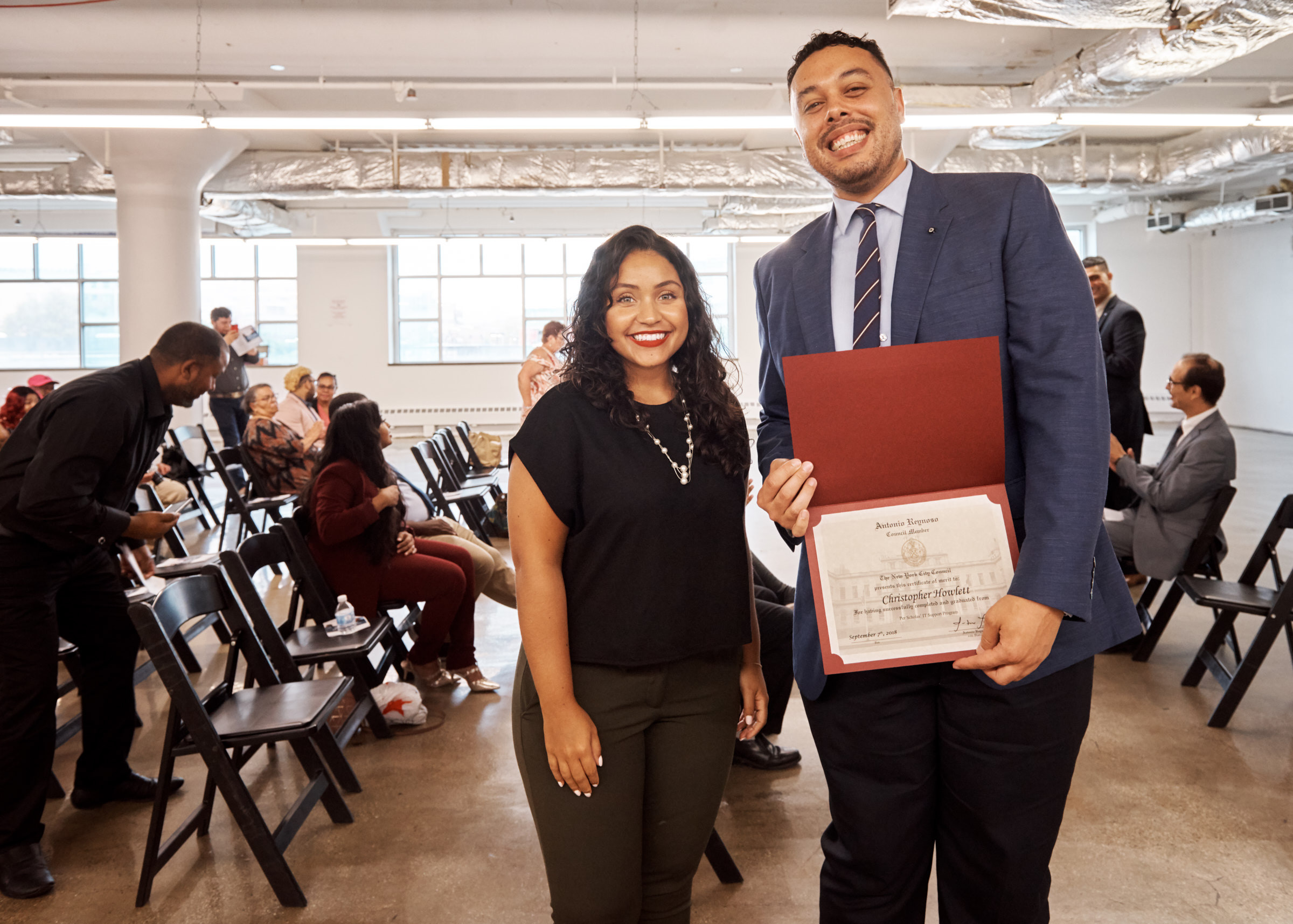 Two smiling adults stand in a room of people; the man holds up a certificate folder.