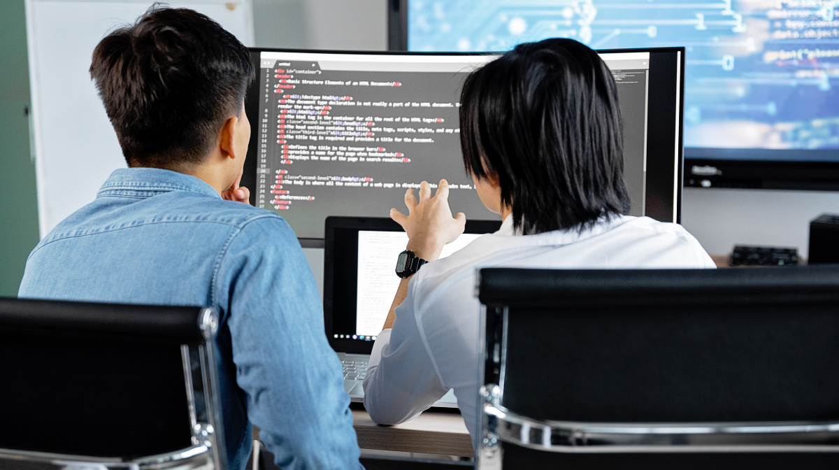 Two young professionals sit at a desk reviewing code on a large monitor, one person pointing at the screen while the other listens.