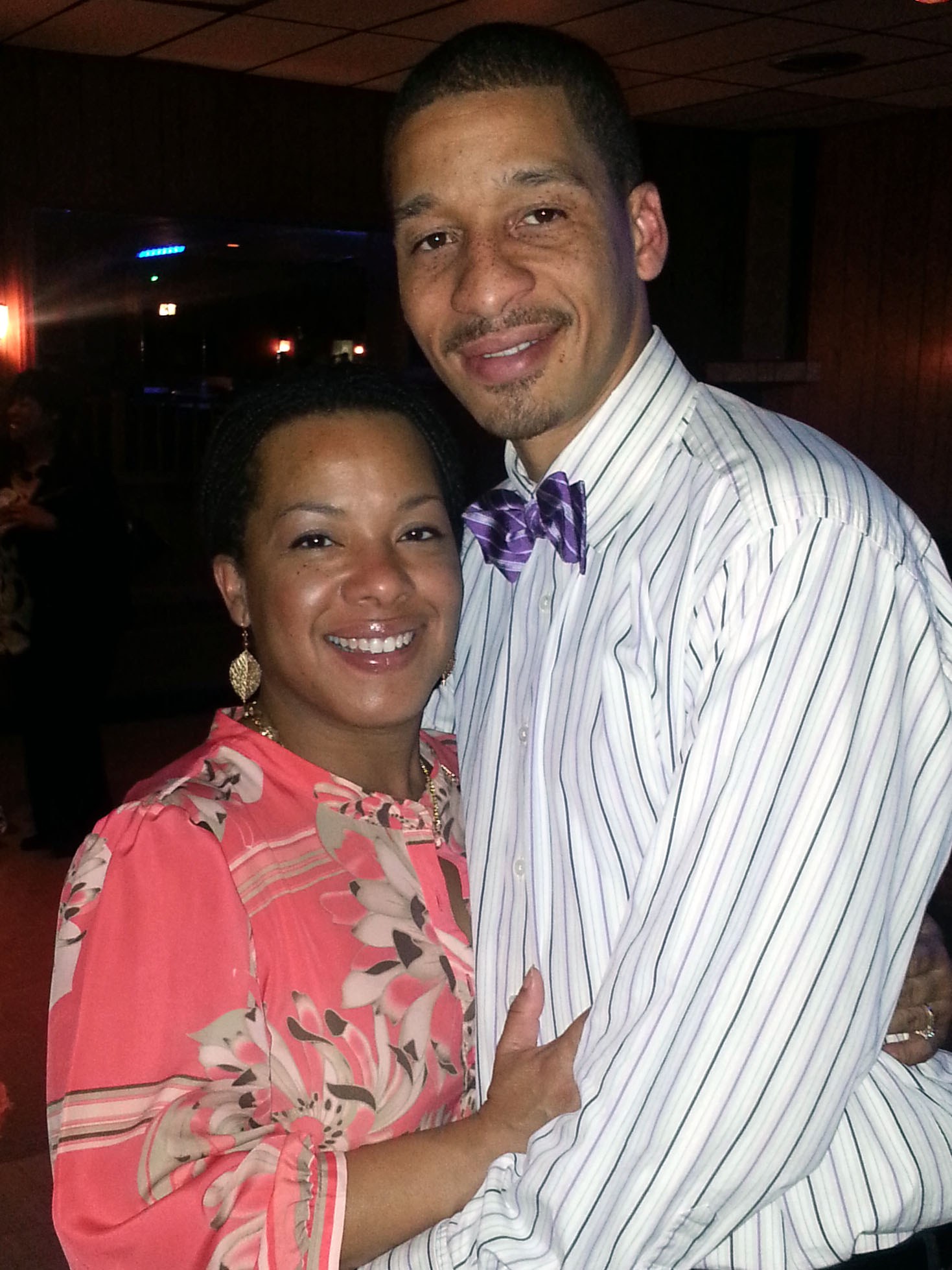 A smiling adult couple embracing and looking at the camera in an indoor event setting.