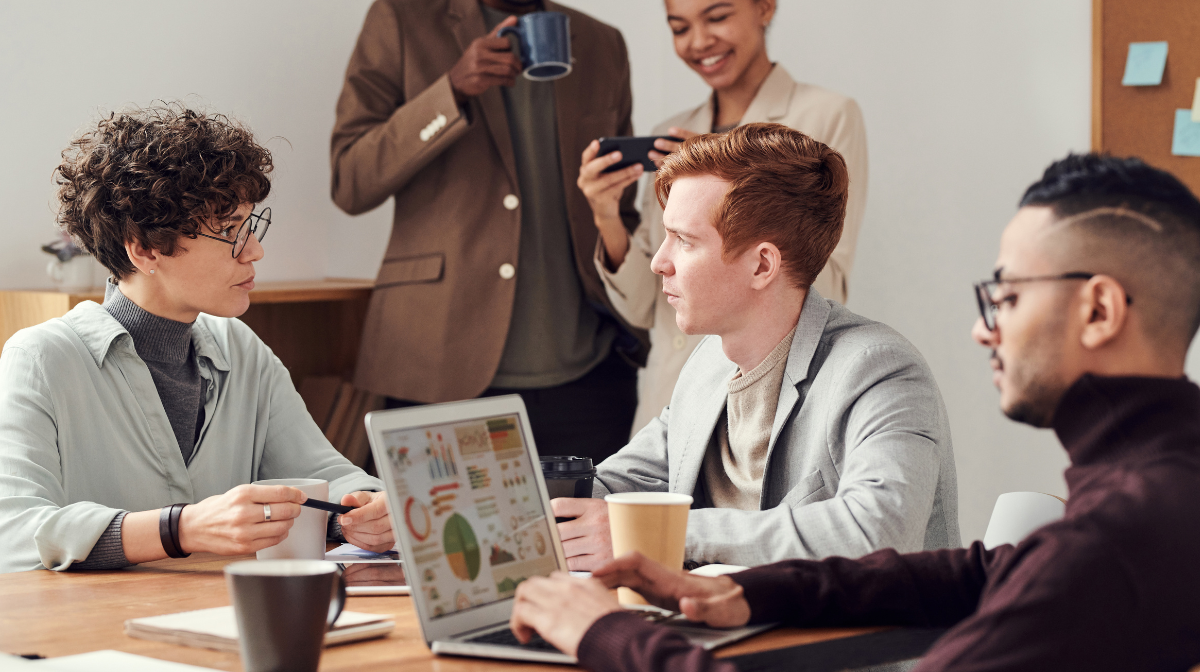 A group of colleagues sit around a table in discussion, with a laptop displaying charts and colleagues sharing a laugh together in the background.
