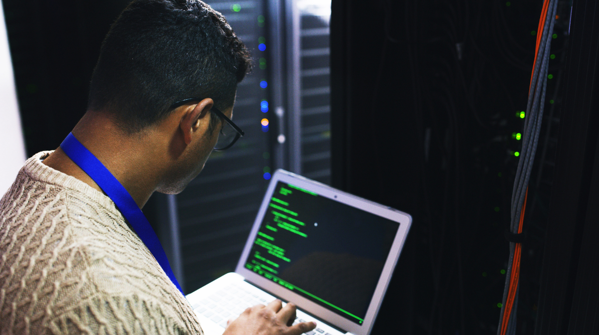 Man working on a laptop with code in a server room.