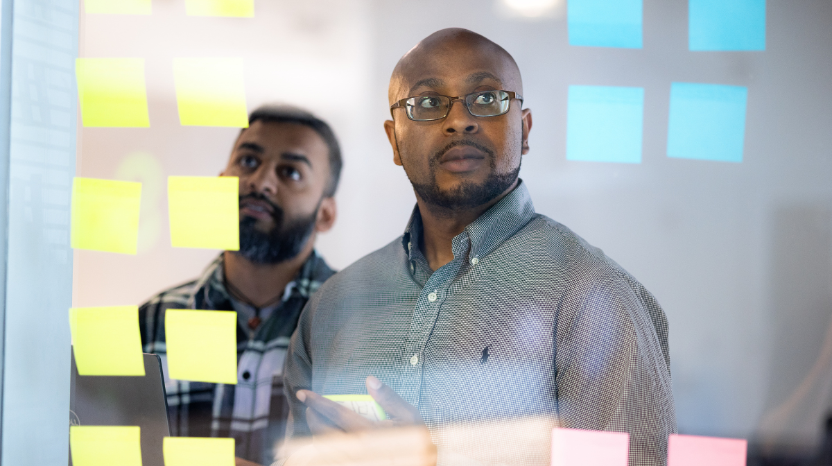 Two men colleagues standing behind a glass wall covered in sticky notes, looking thoughtfully at the notes.