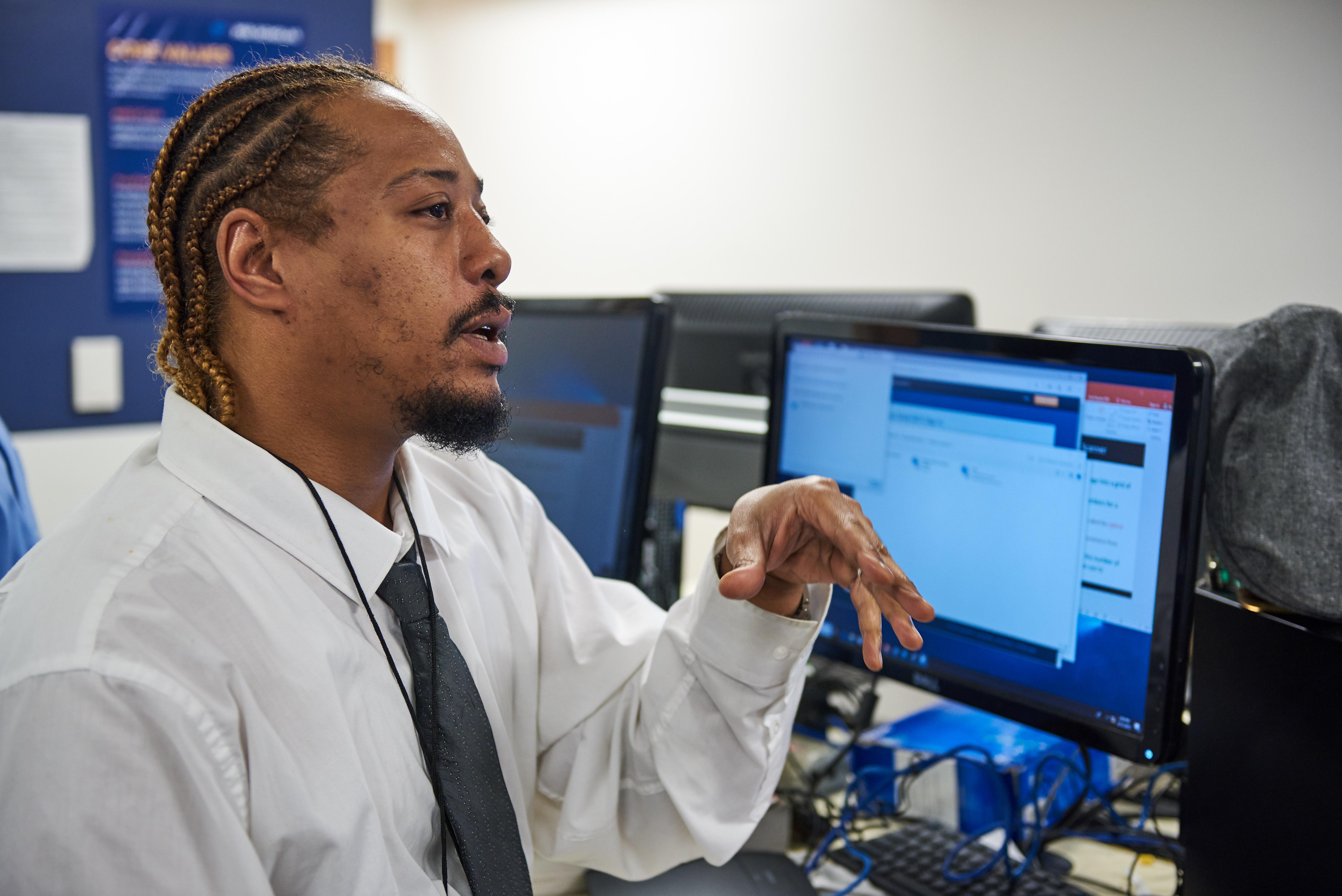 Man speaking while seated at a computer workstation in a classroom.