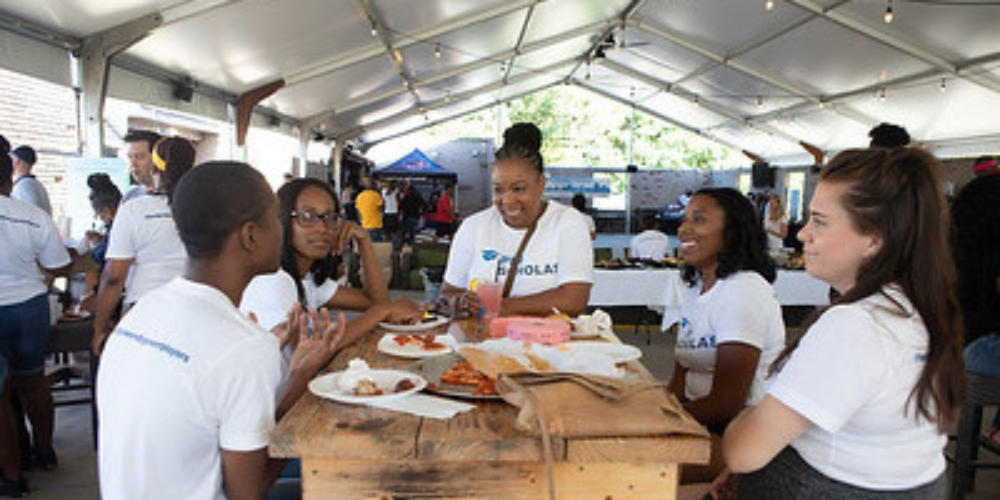Five Per Scholas students sit around a picnic table under a large tent, smiling and talking across plates of food.