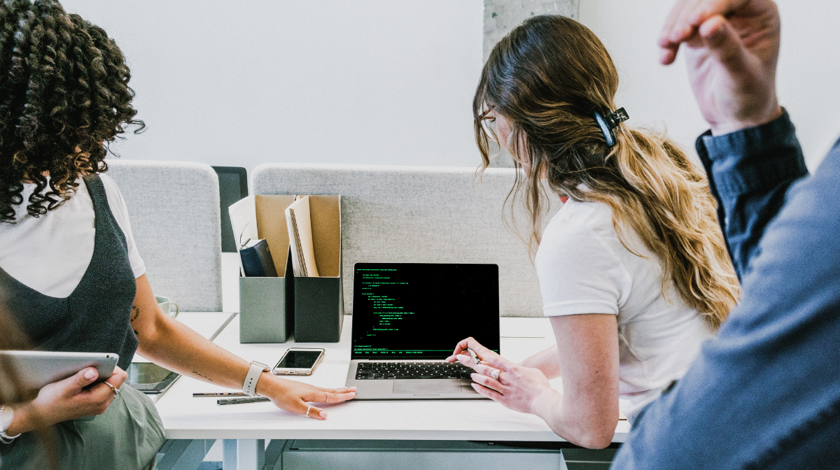 Three young professionals collaborating at a desk with a laptop displaying code.