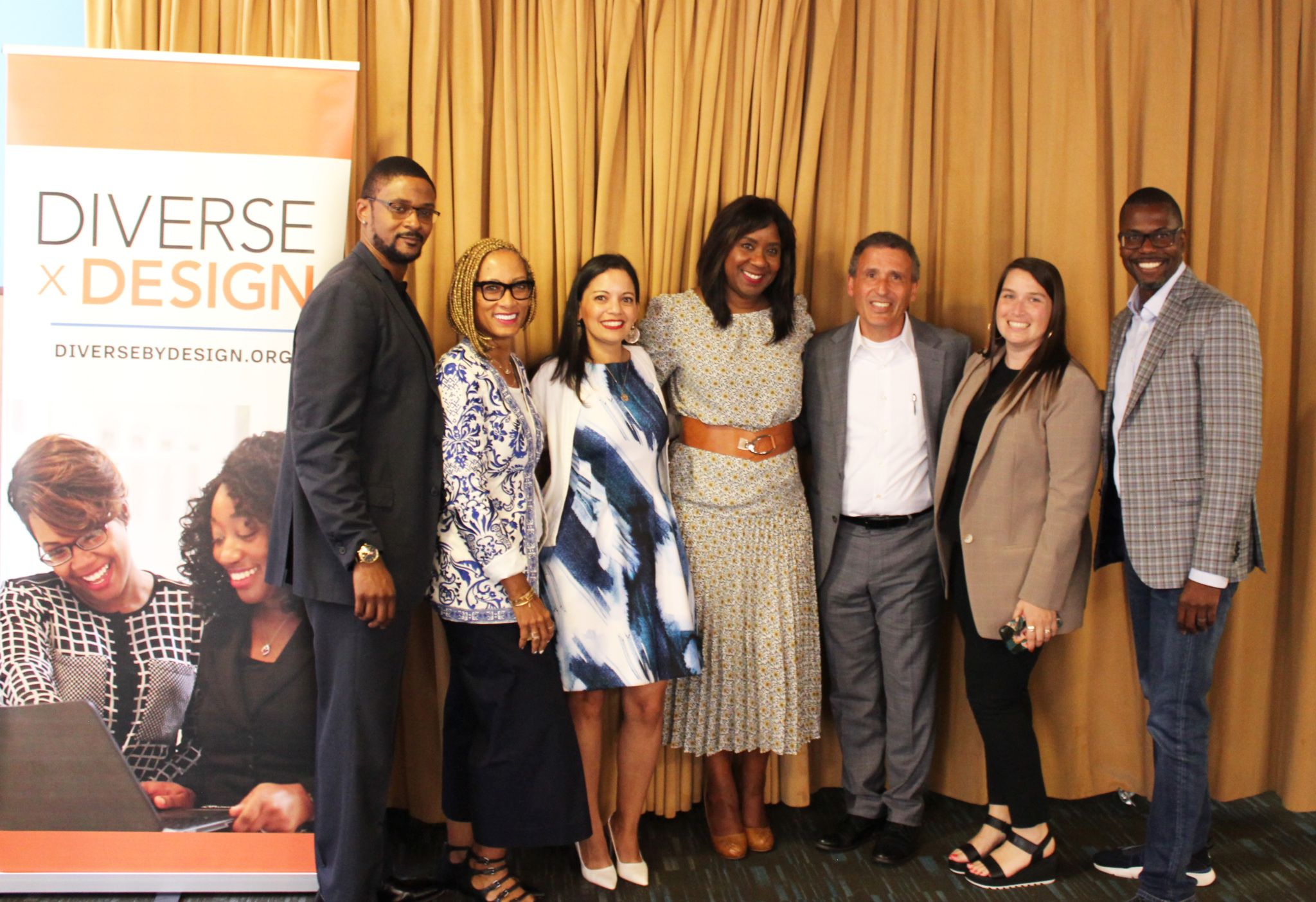 Seven professionally dressed adults stand smiling in a row at a Diverse by Design event banner.