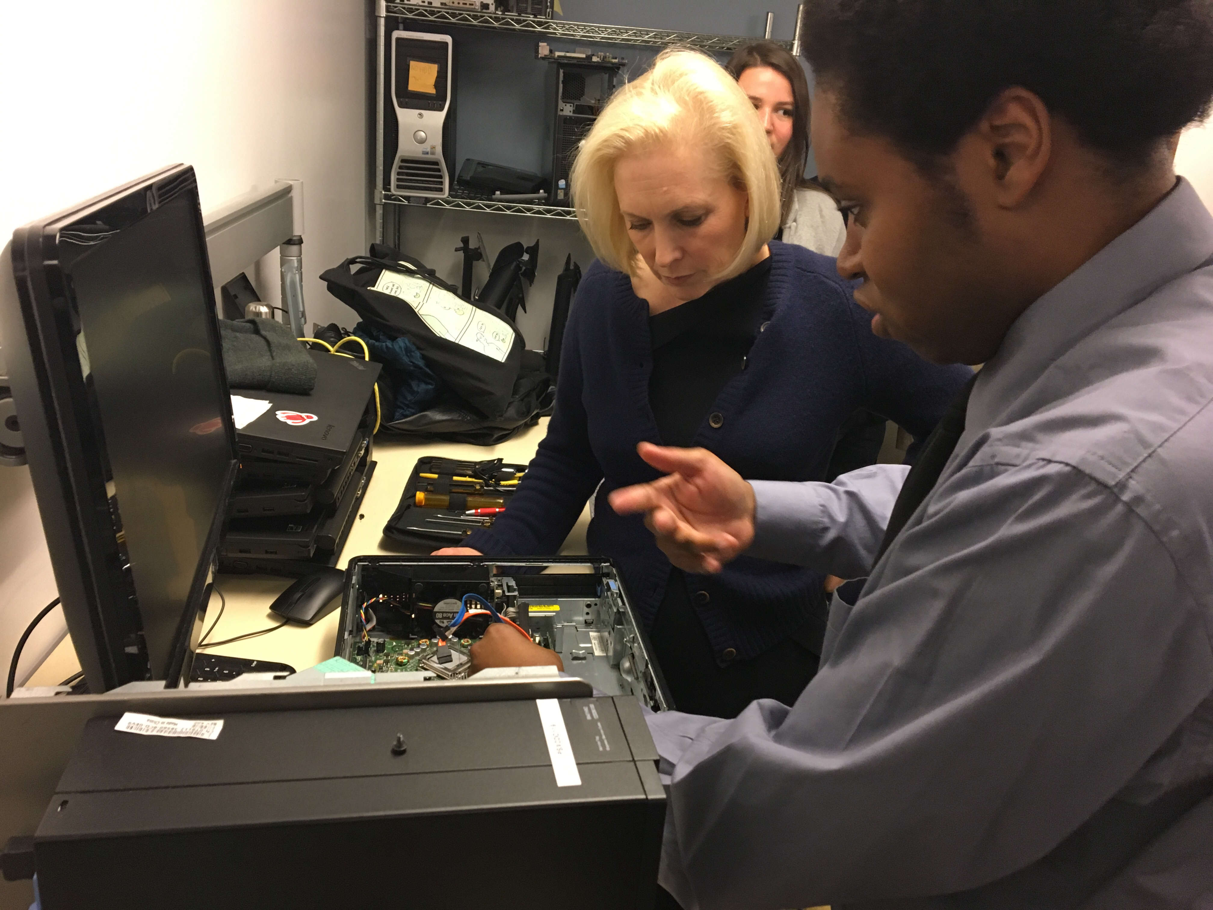 A visitor watches as a technician explains the components inside an open desktop computer on a workbench.