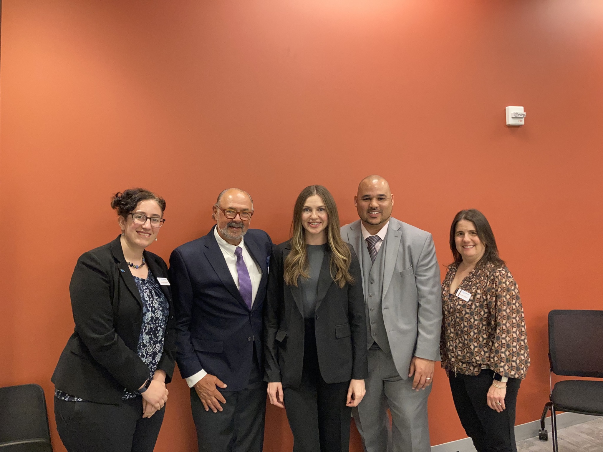Five staff and partners stand shoulder-to-shoulder smiling in front of an orange wall, posing for a group photo.