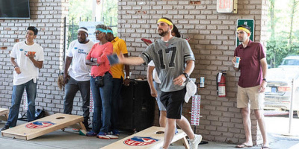 Adults wearing headbands play cornhole while others watch in an indoor space.