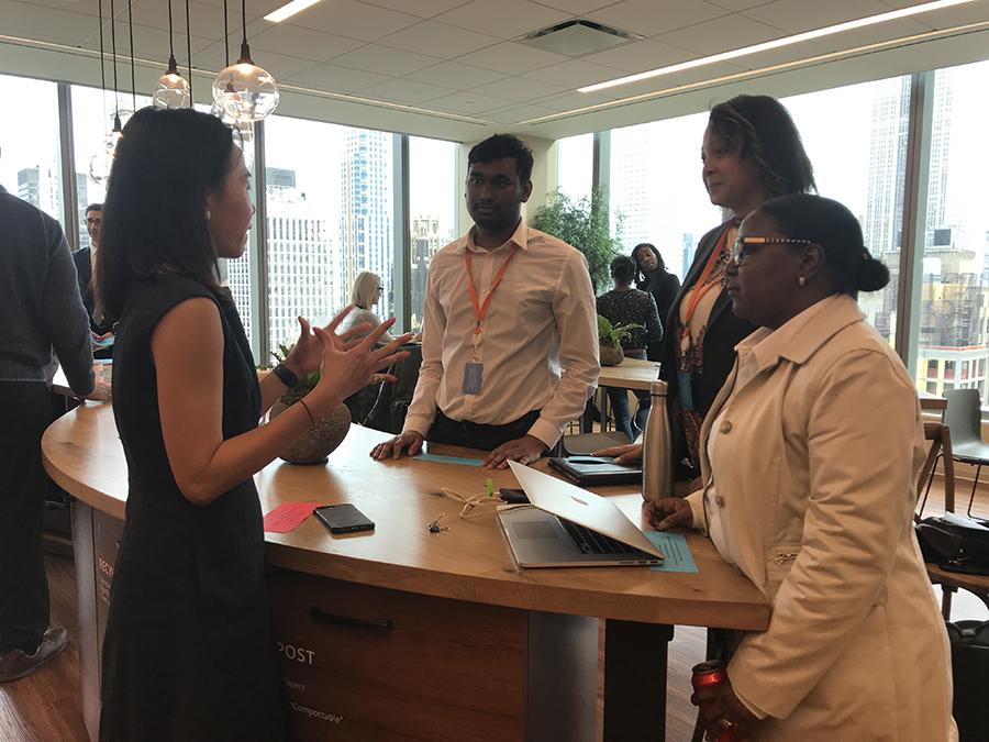 A woman speaks and gestures while three adults listen around a workspace with a laptop.