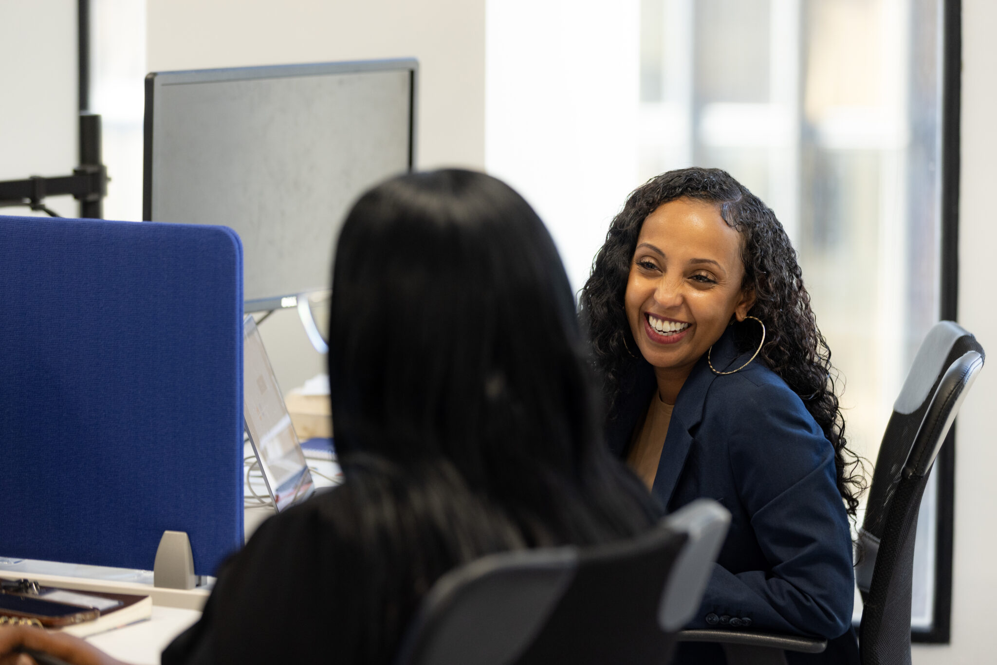 Two women smiling and talking at adjacent desks in an office setting.