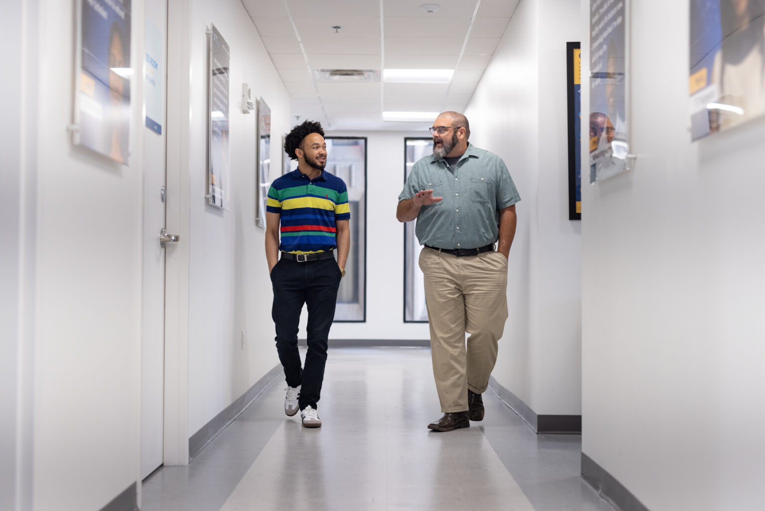 Young man in a striped polo shirt smiling and talking with an older man in a denim shirt as they walk down a hallway.