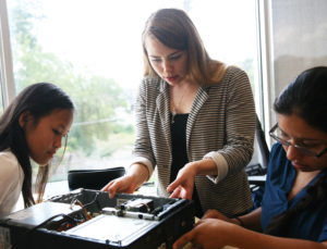Instructor guiding two students as they assemble computer hardware in a classroom.