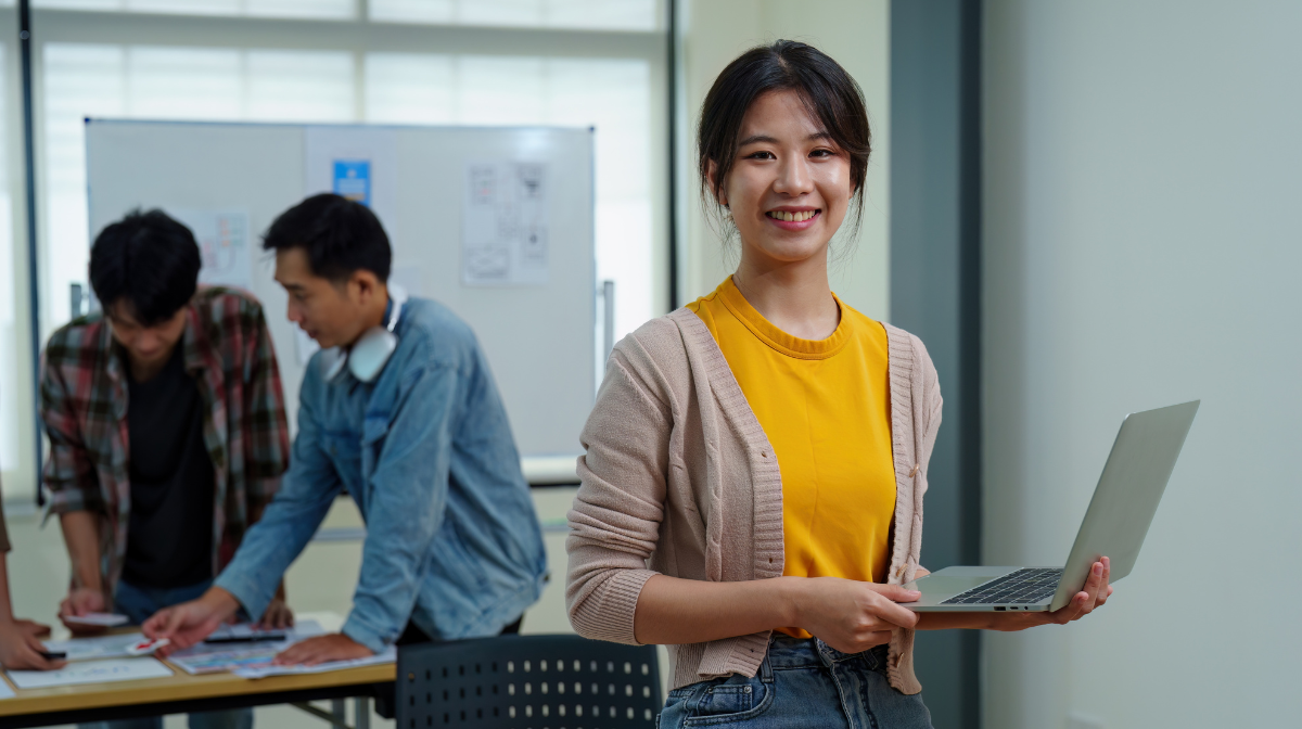 Female learner smiling and holding a laptop in a classroom while classmates collaborate at a table behind her.