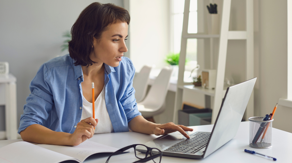 Woman working at a desk, looking at a laptop screen while holding a pencil and taking notes.