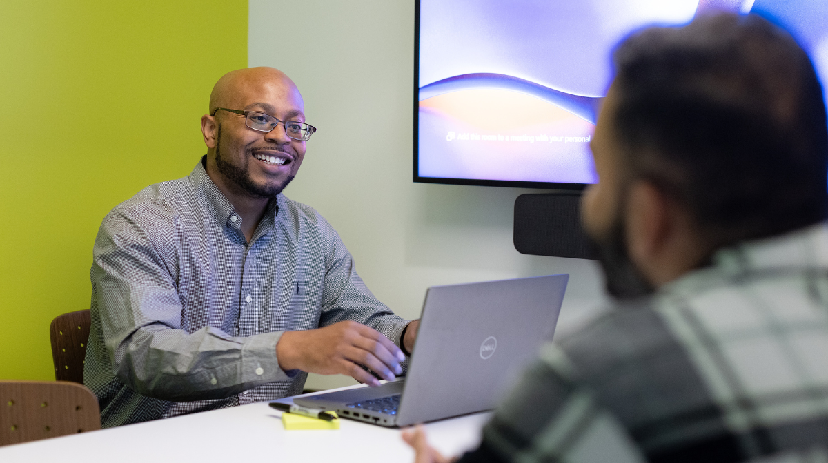 Two male learners having a friendly meeting at a table with a laptop and screen in a small conference room.