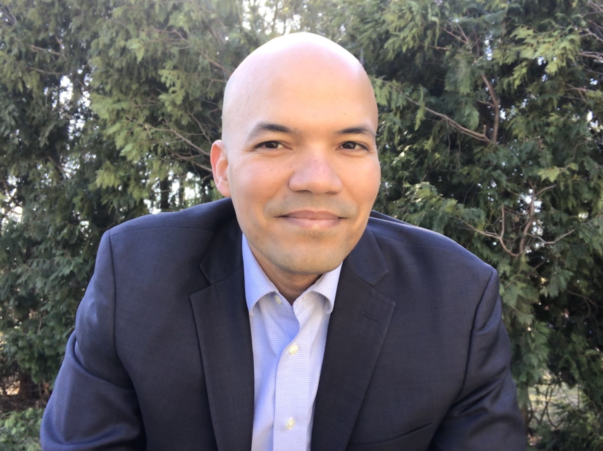 Man in a suit smiling outdoors with trees behind him.