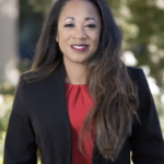 Woman smiling confidently in professional attire, standing outdoors with blurred trees behind her.