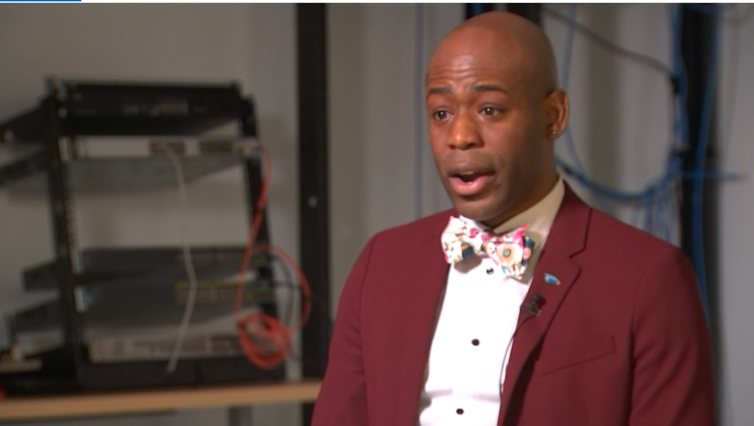 Man speaking in an interview in a tech workspace with networking equipment and cables visible behind him.