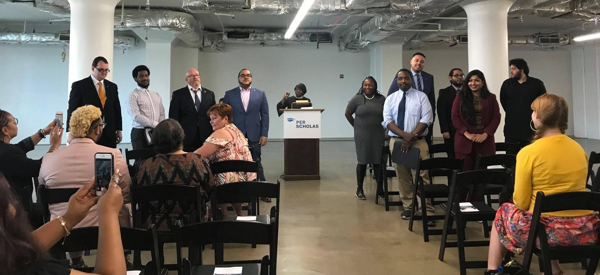 Per Scholas staff and graduates standing at front of a room near a podium with an audience taking photos.