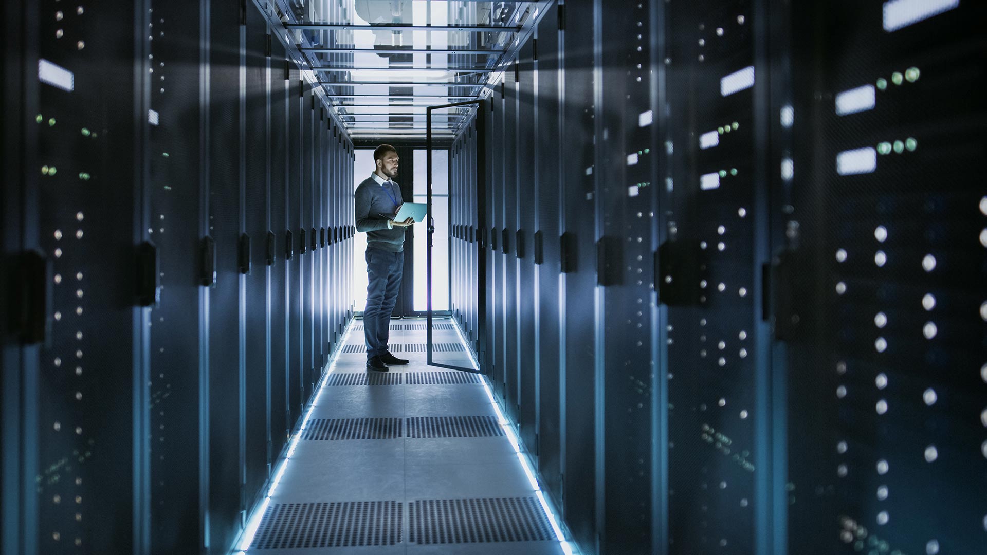 Man standing in a server room aisle holding a tablet, inspecting racks of servers.