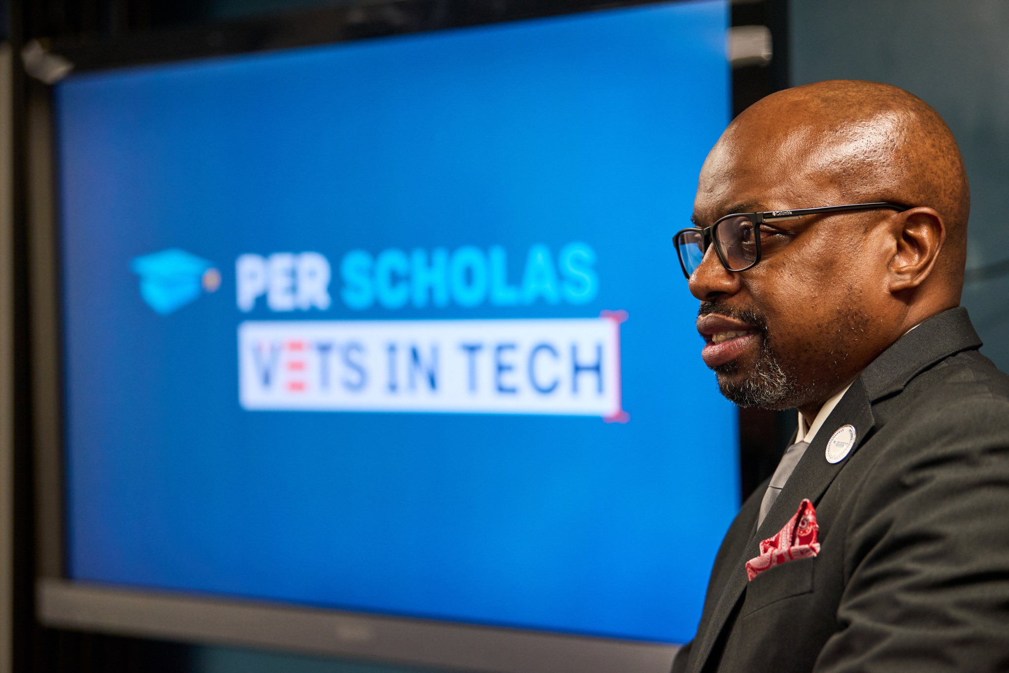 Man speaking in front of a screen displaying the Per Scholas Vets in Tech program logo.