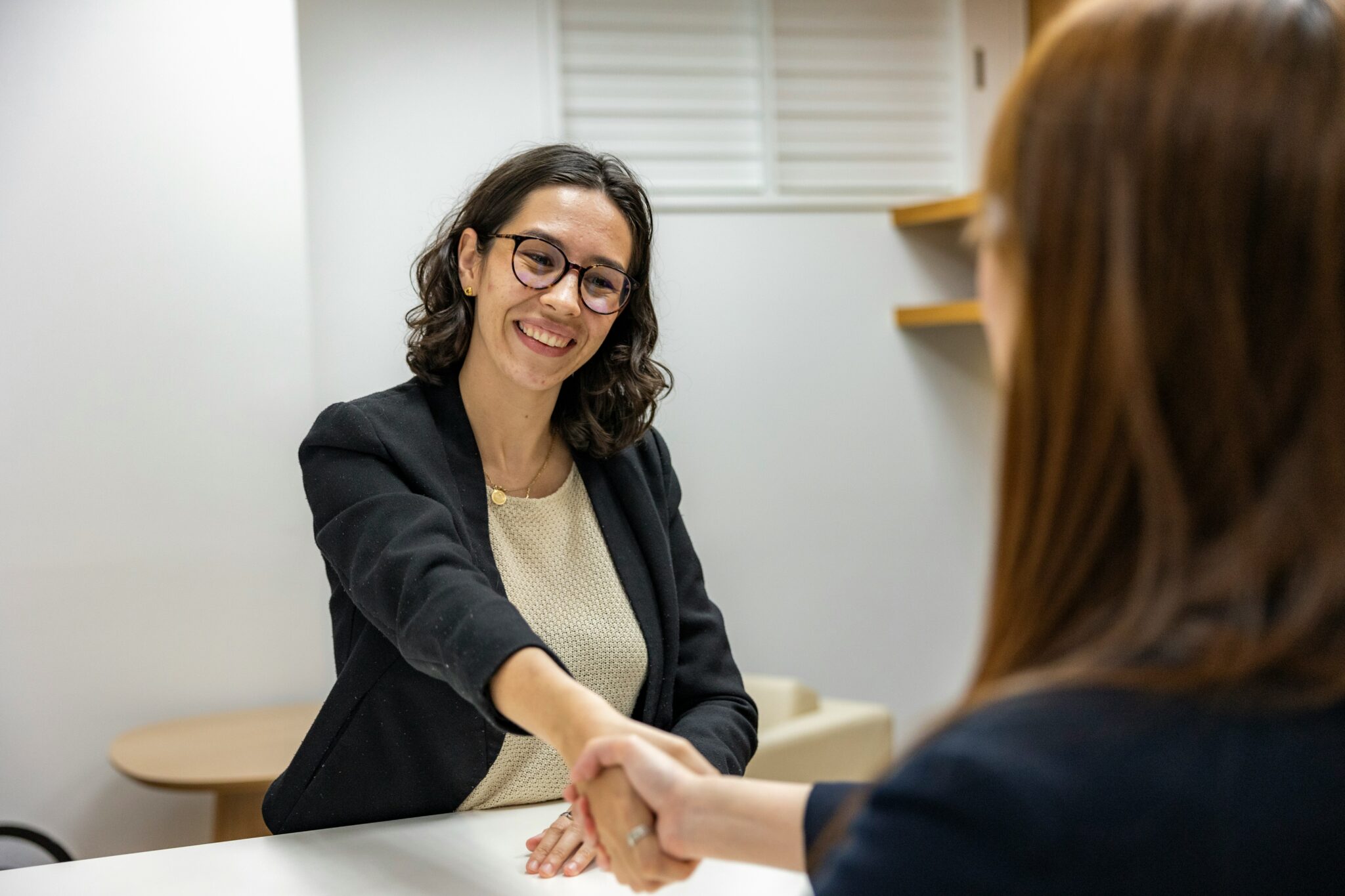 Smiling young female professional with curly hair and glasses shaking hands with a colleague across a table in a bright office setting.